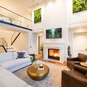 Bright, modern living room with high ceilings, white walls, a sectional sofa, two brown leather chairs, round gold coffee table, fireplace, wall-mounted TV, plants, and large green art pieces above the mantel.