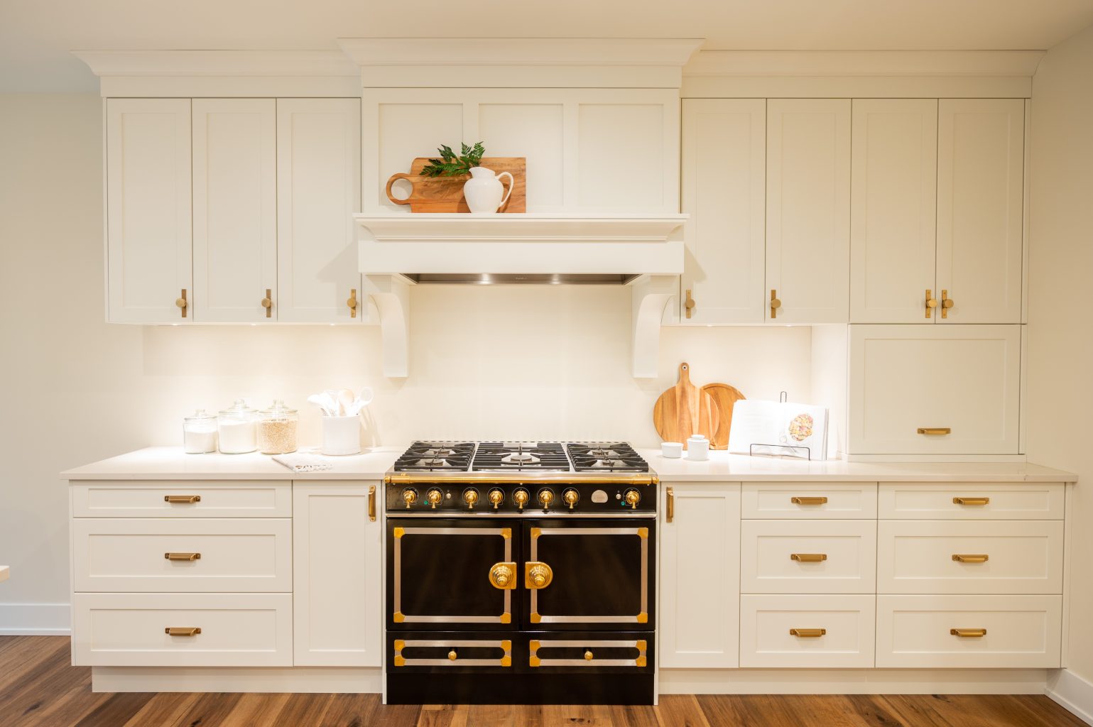 A modern kitchen with white cabinets, gold handles, a black and gold stove, wooden cutting boards, and minimalist decor on white countertops and shelves. Warm lighting highlights the clean, elegant design.