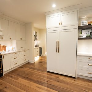 Modern kitchen with white cabinets, brass handles, a black and gold stove, wooden cutting boards, open shelves with plants and photos, and wide wooden floors under warm lighting.