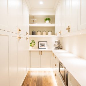 Bright, modern pantry with white cabinets, gold handles, open shelves holding jars and plants, wooden floor, a mixer, and a microwave on a marble countertop under soft recessed lighting.