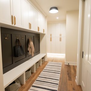 A modern mudroom with white cabinets, gold handles, a striped rug, wooden floor, hooks with hanging bags and coat, and storage baskets under a built-in bench. Bright overhead lighting illuminates the space.