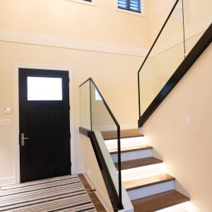 A modern entryway with a black front door, wooden stairs with glass railing, steps illuminated by built-in lights, beige walls, high windows with blinds, and a striped rug on the floor.