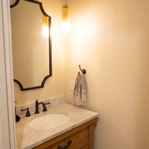 A small bathroom with a wooden vanity, white countertop, oval sink, black faucet, a scalloped-edge mirror, pendant light, and a hand towel hanging on the wall. Cream-colored walls create a warm atmosphere.