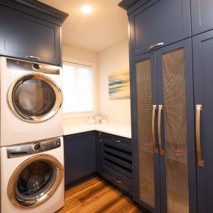 A modern laundry room with stacked washer and dryer, blue cabinets with gold handles, wood flooring, and a window with white blinds. A blurry painting hangs on the wall above the countertop.