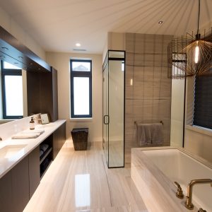 Modern bathroom with a freestanding tub, glass shower, double sink vanity, gold fixtures, large windows, dark cabinetry, and a unique geometric pendant light. Light floor tiles and neutral tones create a clean, elegant look.