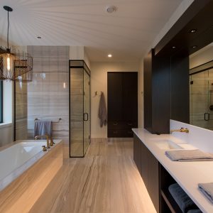 Modern bathroom with a large soaking tub, spacious glass shower, double sink vanity with mirrors, dark wood cabinets, gold fixtures, folded towels, and a hanging decorative light fixture. Natural light streams in from big windows.