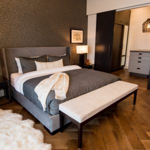 Modern bedroom with a gray upholstered bed, dark bedding, and a cream bench at the foot. Wooden floors, fur rug, nightstands with lamps, and a walk-in closet with dark cabinets and a pendant light are visible.