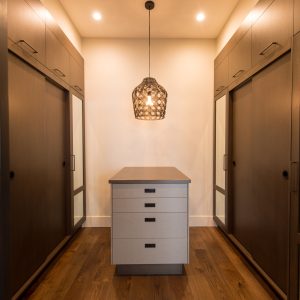 A modern walk-in closet with dark wood cabinets, a central white island with drawers, wood flooring, and a woven pendant light hanging from the ceiling. Mirrors are mounted on the cabinet doors.