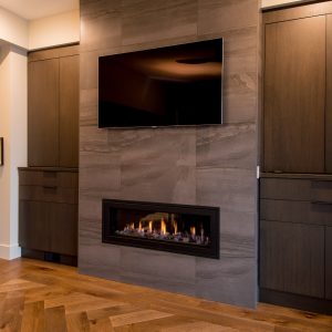 A modern living room features a wall-mounted TV above a sleek built-in gas fireplace, flanked by dark wood cabinets, with light brown herringbone wood flooring and soft, warm lighting.