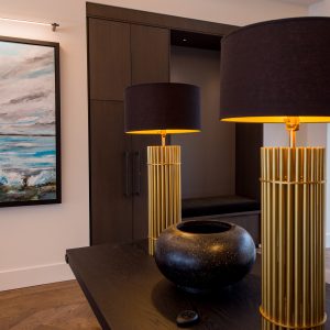 A modern room features a large painting of the ocean on the wall, a black table with a round black vase, and a gold lamp with a black shade reflected in a mirror. A window shows greenery and a pool outside.