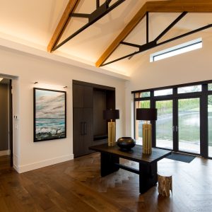 Modern entryway with vaulted ceiling, exposed beams, large glass doors, a wooden floor, table with two lamps, and a landscape painting on the wall; a cat is partially visible near the table legs.