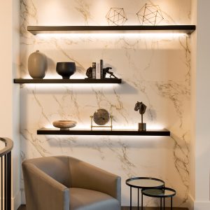 A modern reading nook with a gray armchair, two round side tables, and three floating shelves displaying sculptures, vases, books, and geometric decor against a marble wall, all softly lit with built-in lighting.