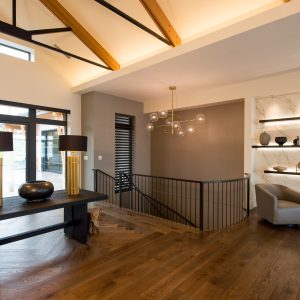 A modern interior with wooden floors, a dark table with gold lamps, a gray armchair, floating shelves with decor, a metal railing, exposed beams, large windows, and a contemporary chandelier.