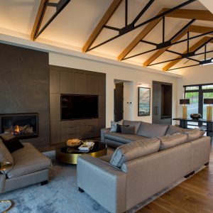 Modern living room with exposed wooden ceiling beams, large windows, gray sofas, a round coffee table, a wall-mounted TV, and a black fireplace. Natural light brightens the open and spacious area.