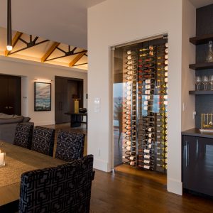 Modern dining room with a long wooden table, patterned chairs, hanging pendant lights, a glass wine display, shelves with glasses, and a view into a living area with contemporary decor.