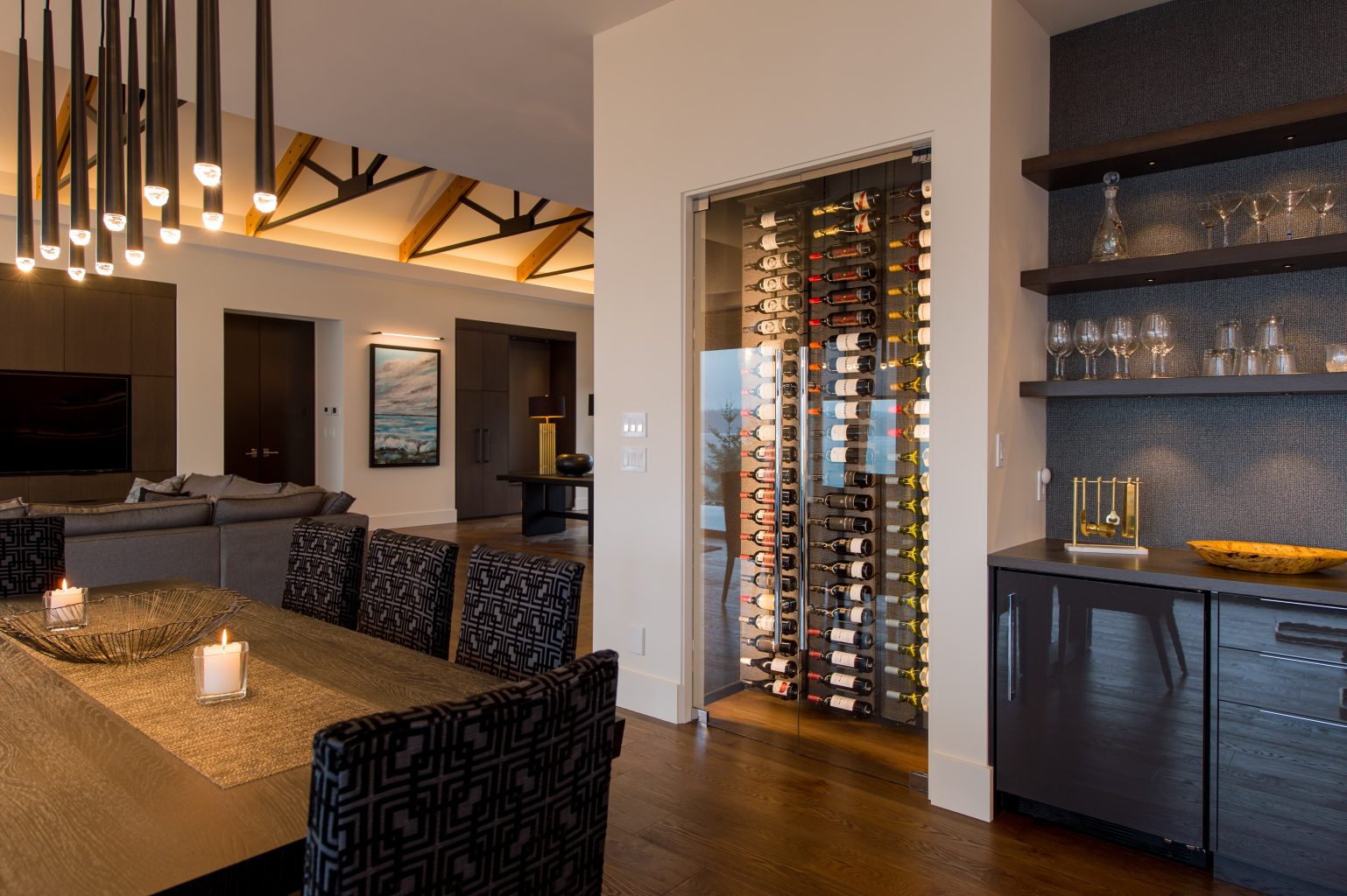 Modern dining room with a long wooden table, patterned chairs, lit candles, and a glass-enclosed wine rack. Contemporary lighting and exposed ceiling beams complete the open-concept space, blending into a living area.