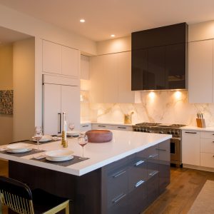 Modern kitchen with white cabinets, marble backsplash, a large island with dark drawers, two place settings, gold-accented chairs, and under-cabinet lighting. Warm wood flooring and stainless steel appliances.