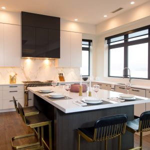A modern kitchen with white cabinets, marble backsplash, large island with place settings, black and gold bar stools, dark wood floors, and large windows letting in natural light.