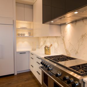 Modern kitchen with white cabinets, marble backsplash, stainless steel stove and range hood, wooden floor, open shelves displaying dishes, and soft under-cabinet lighting.