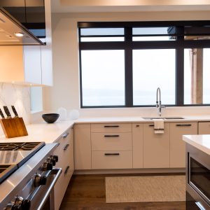 Modern kitchen with white cabinets, a large window above a sink, a gas stove, knives in a wooden block, a bowl, a microwave on an island, and wood flooring. Natural light fills the space.