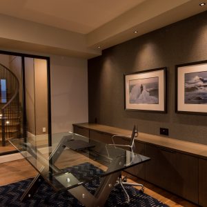 Modern home office with a glass desk, metal chair, and dark wood cabinets. Two framed landscape photos are on the wall, and a glass door reveals a spiral staircase and soft lighting beyond.