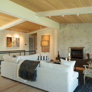 A modern living room with a light wood ceiling and floor, white sectional sofa, black throw, and armchairs. There’s a brick fireplace, artworks on the wall, and large windows bringing in natural light.