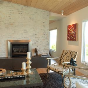 A modern living room with two patterned chairs, a glass coffee table with candles and decor, a gas fireplace with a brick surround, and large windows letting in natural light. Bright abstract art hangs on the wall.
