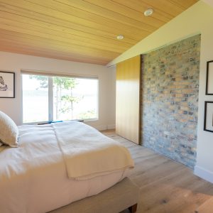 A bright, modern bedroom with a large window, wood ceiling, light bedding, a brick accent wall, framed artwork, and sliding wooden closet door. Natural light fills the room, highlighting minimalist decor.