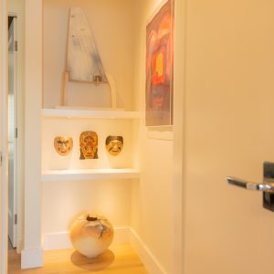 A small, well-lit alcove features three sculpted face masks on a white shelf, a large decorative vase on the floor, abstract artwork on the wall, and light wood flooring with a wood-paneled ceiling above.