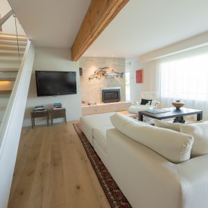 A modern living room with light wood floors, a white sectional sofa, a flat-screen TV, a fireplace, and large windows with sheer curtains. A wooden beam and a staircase with glass railing are visible on the left.