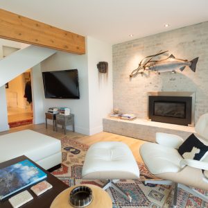 Modern living room with a white lounge chair and ottoman, wall-mounted TV, fireplace set in a brick wall, large fish sculpture above the mantel, and a patterned rug on light wood floors.
