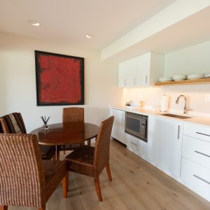 A modern kitchenette with white cabinets and countertop, a sink, microwave, and open shelves with dishes. A round wooden dining table with four wicker chairs sits below a large red abstract painting.