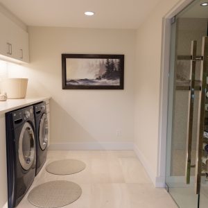 A modern laundry room with two front-loading machines, white cabinets, a countertop with seashells, a basket, a framed painting, and a glass wine cellar filled with bottles on the right.