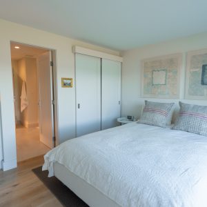 A bright, modern bedroom with a white bed, two patterned pillows, white drawers, artwork above the bed, a cross on the wall, and an open door leading to a well-lit hallway or bathroom.