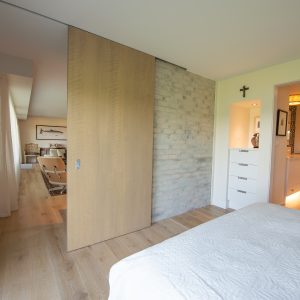 A modern bedroom with light wood floors and a white bedspread, featuring a large wooden sliding door partially open to a living area. The room has neutral walls and a doorway leading to a bright bathroom.