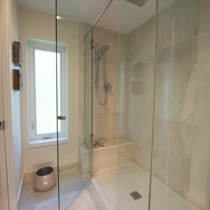 A modern bathroom with a glass-enclosed shower, beige tiles, a built-in bench, rain showerhead, frosted window, towel hooks, and a small basket on the floor.