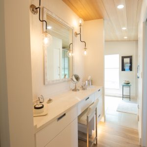 A bright, modern vanity area with a white countertop, a wall mirror, two globe light fixtures, and a cushioned stool. Light wood floors and ceiling add warmth to the space, extending into a sunlit hallway.