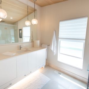 Modern bathroom with a white vanity, large mirror, hanging pendant lights, towel on a rack, window with a blind, framed artwork, glass side table, and soft natural lighting.