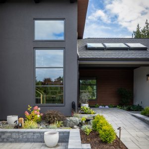 Modern house exterior with dark gray and wood siding, large windows, and a landscaped front yard featuring paved walkways, a raised garden bed, small shrubs, rocks, and potted plants under a partly cloudy sky.