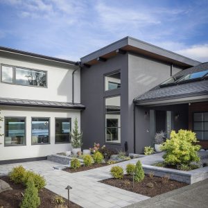 Modern two-story house with white and dark gray exterior, large windows, and a landscaped front yard with shrubs and pavers under a blue sky with scattered clouds.