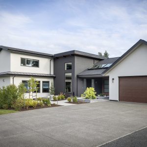 Modern two-story house with white and gray exterior, large windows, and wooden garage door. The home has landscaped plants in front and overlooks a body of water with trees in the background.
