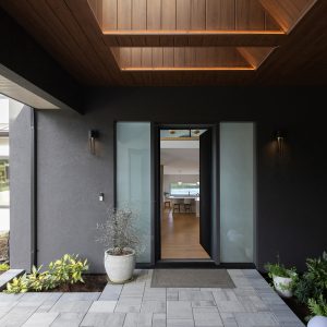 Modern home entrance with dark walls, a large glass door, potted plants, rectangular gray tiles, and a wooden ceiling featuring a rectangular skylight. The entryway leads to a bright, open interior space.