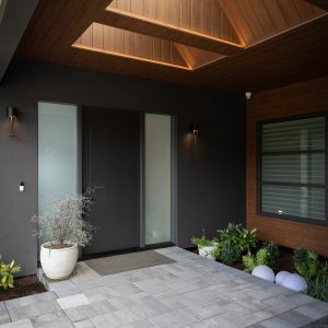 Modern house entrance with a large, dark door, frosted glass panels, potted plants, and overhead wooden ceiling featuring skylights. The porch has gray stone tiles and minimalist exterior lighting.