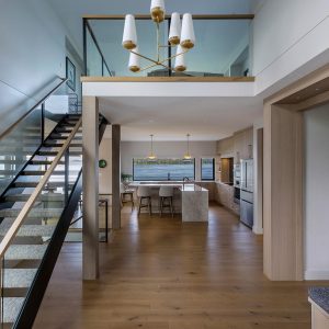 Modern open-concept kitchen and dining area with wood floors, light wood cabinetry, a white island with bar stools, a staircase with glass railing, and a loft above with a contemporary chandelier.