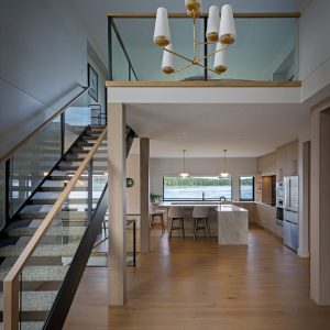 Modern open-concept kitchen with light wood floors, a marble island, barstools, staircase with glass railing, and a gold chandelier. Large windows provide natural light and a view of greenery outside.
