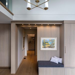 A modern entryway with light wood walls and floors, built-in bench with cushions, framed landscape artwork, recessed lighting, and a gold chandelier with white shades hanging above.