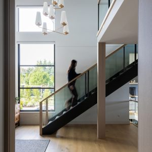A blurred figure walks up a modern staircase with glass railings in a bright, open living space featuring large windows, wooden floors, and a white chandelier.