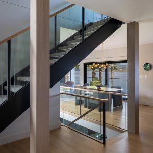 A modern interior with a wooden staircase featuring glass railings, light wood floors, and an open dining area with large windows and a contemporary chandelier.
