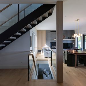 A modern interior with an open staircase featuring glass railings, light wood floors, and a living room with a sofa, TV, and dining area with hanging lights. Natural light fills the space through large windows.