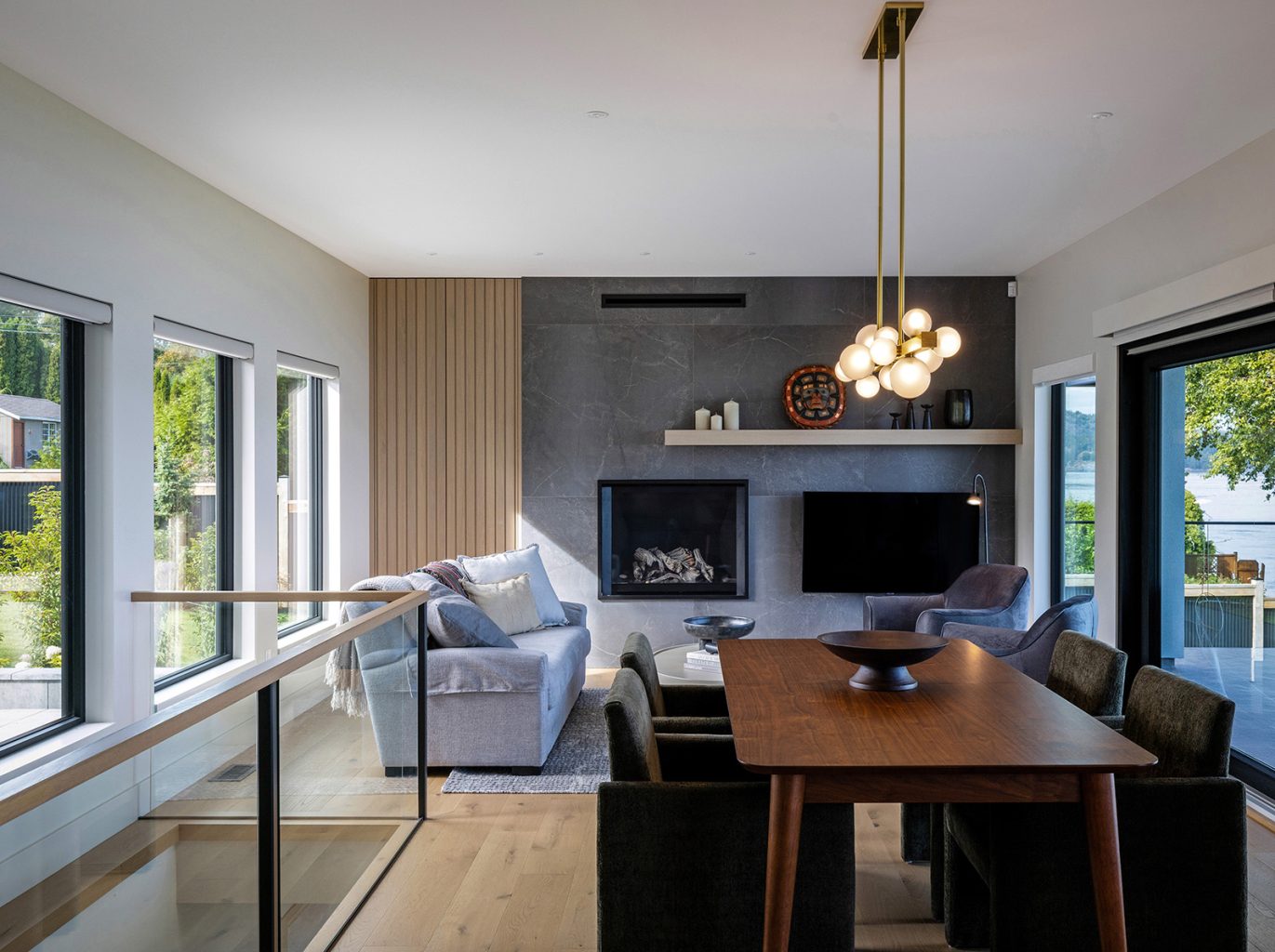 Modern living and dining area with large windows, a wooden dining table, velvet chairs, grey sofas, a fireplace, wall-mounted TV, and stylish pendant lighting. Bright, airy space with views of greenery outside.